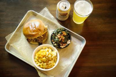 Pulled pork sandwich with mac and cheese, collard greens, and a beer on a metal tray.