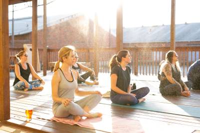 Several women practice yoga outdoors on a wooden deck in sunlight.